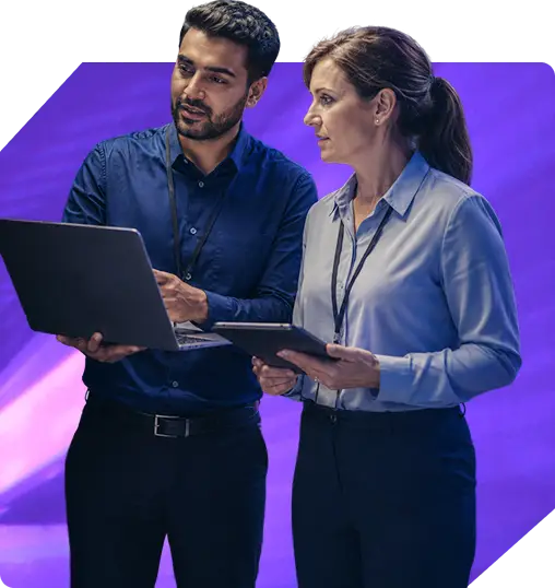 Two professionals, a man and a woman, collaborate on a laptop and tablet in an office setting.